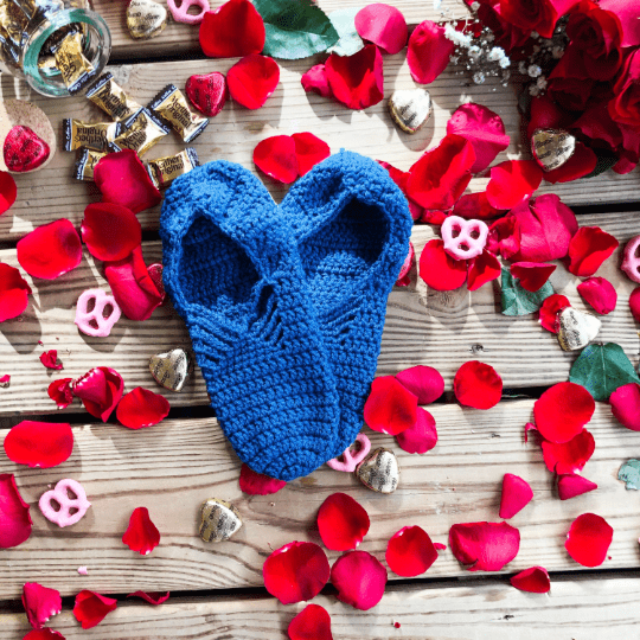 A pair of blue slippers are laid out on a background of wooden slats, rose petals, chocolate hearts, and caramel candies.