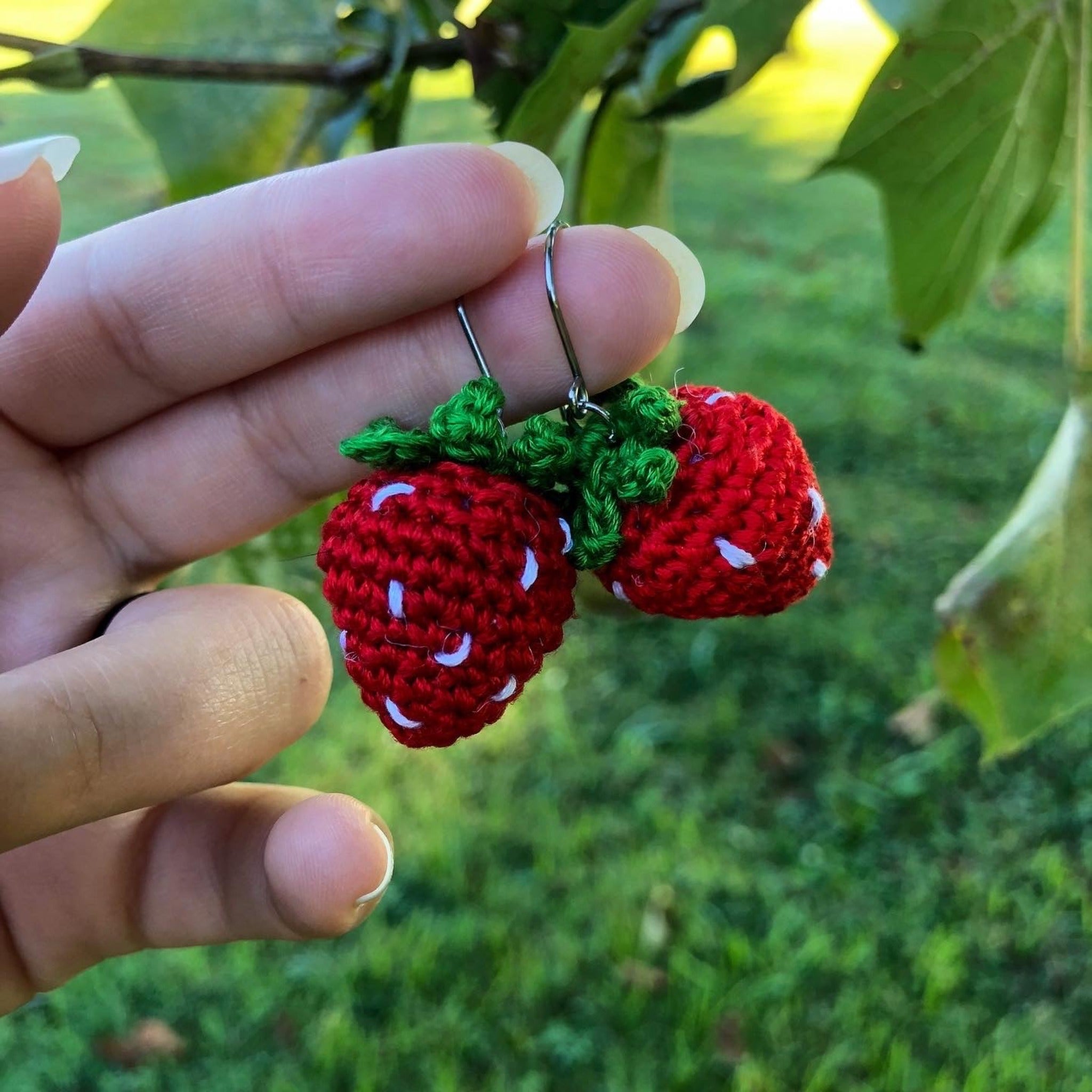 Two red strawberry earrings made out of crocheted embroidery floss are being held on a finger with a green natural background