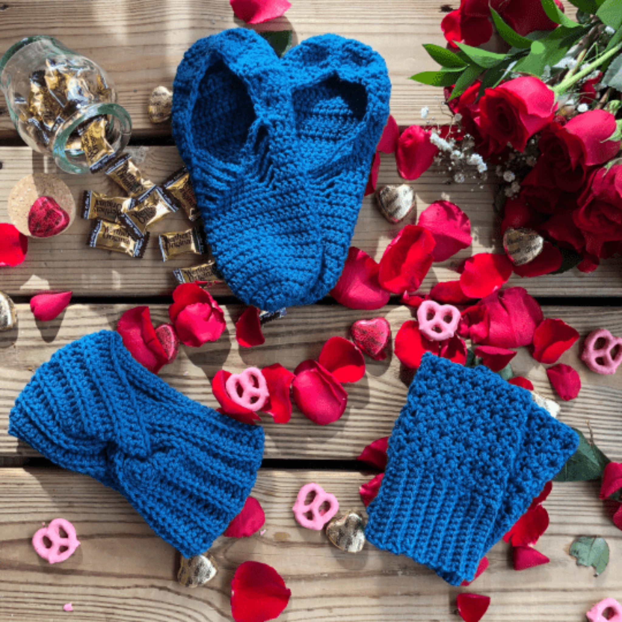 A blue gift set consisting of house slippers, a twisted headband, and fingerless gloves sits on a background of wooden slats, rose petals, pink yogurt covered pretzels, and caramel candies.