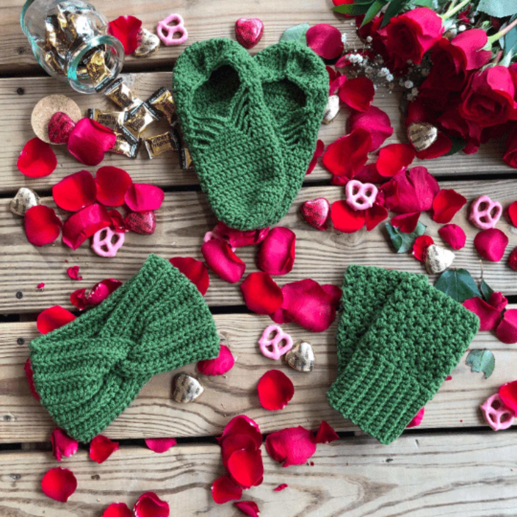 A green gift set consisting of house slippers, a twisted headband, and fingerless gloves sits on a background of wooden slats, rose petals, pink yogurt covered pretzels, and caramel candies.