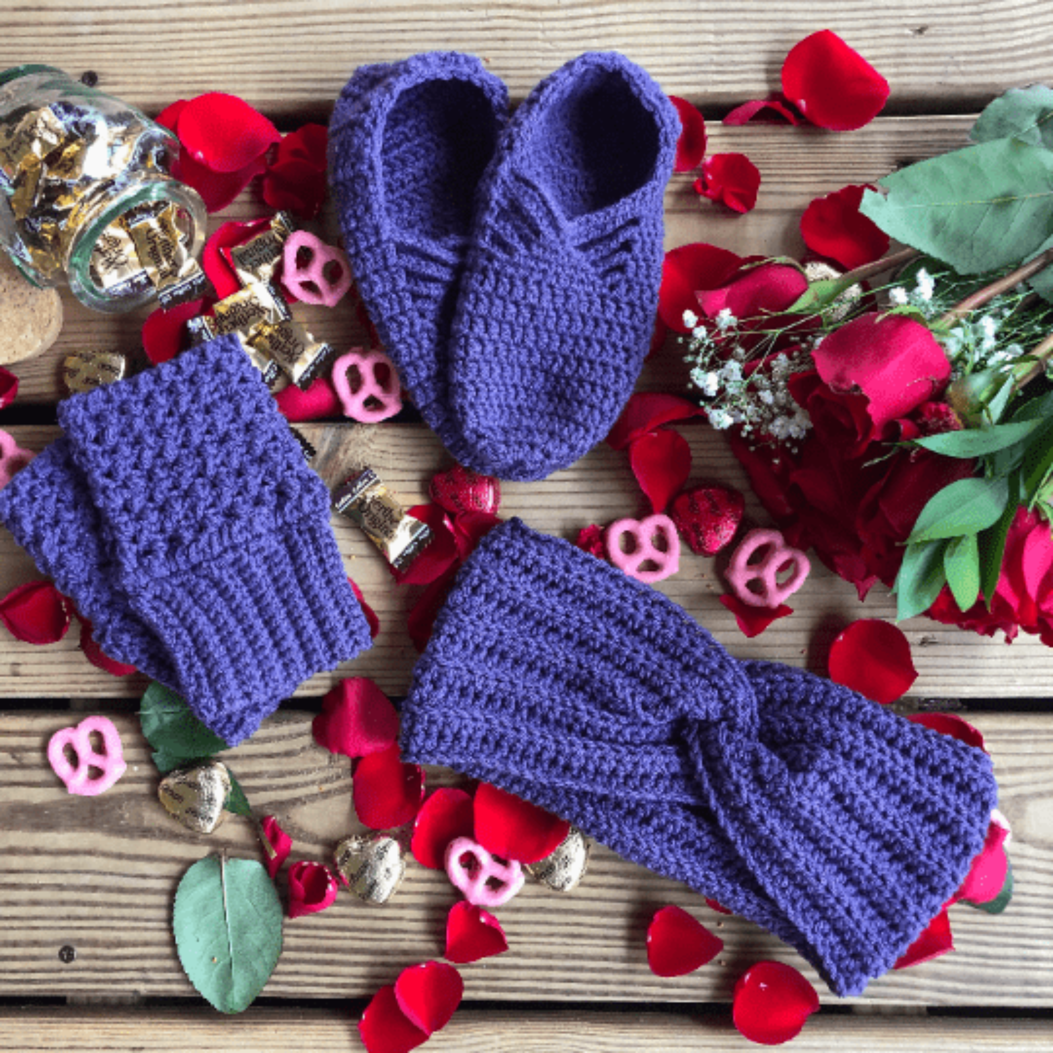 A purple gift set consisting of house slippers, a twisted headband, and fingerless gloves sits on a background of wooden slats, rose petals, pink yogurt covered pretzels, and caramel candies.