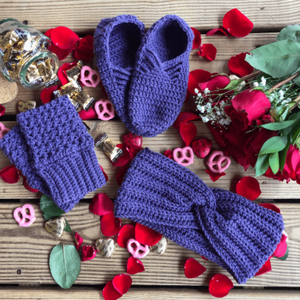 A purple gift set consisting of house slippers, a twisted headband, and fingerless gloves sits on a background of wooden slats, rose petals, pink yogurt covered pretzels, and caramel candies.