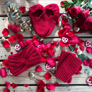 A red gift set consisting of house slippers, a twisted headband, and fingerless gloves sits on a background of wooden slats, rose petals, pink yogurt covered pretzels, and caramel candies.