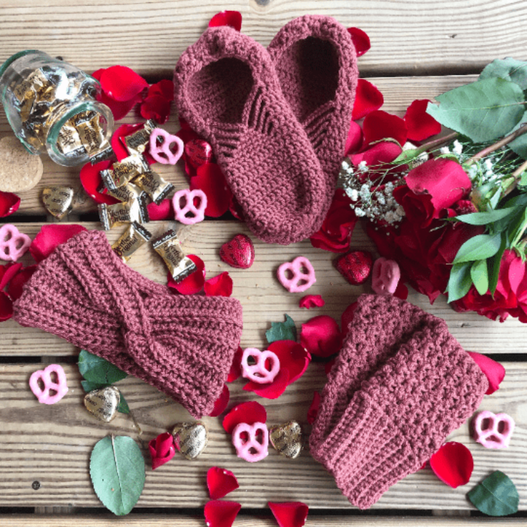 A pink spice gift set consisting of house slippers, a twisted headband, and fingerless gloves sits on a background of wooden slats, rose petals, pink yogurt covered pretzels, and caramel candies.