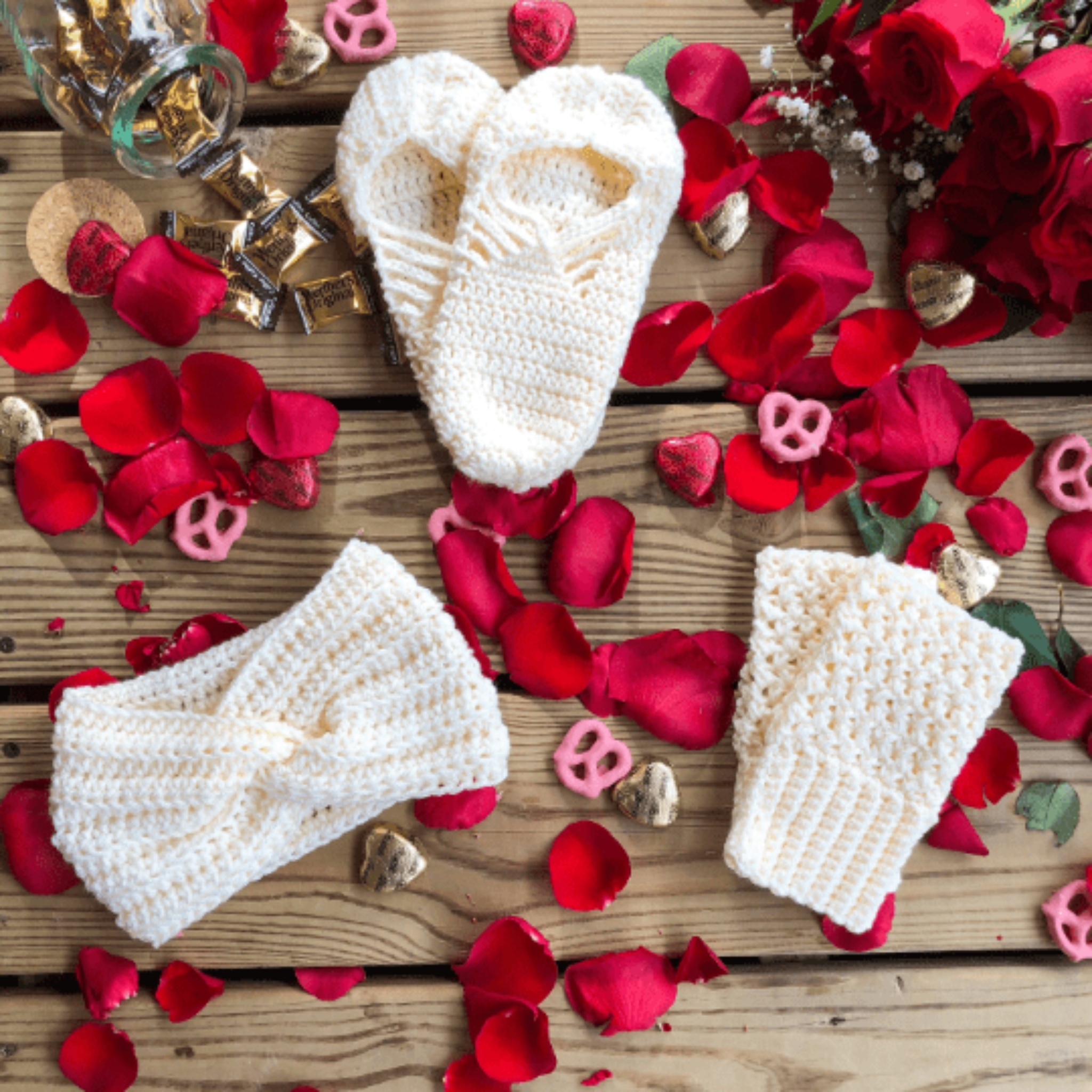 A white gift set consisting of house slippers, a twisted headband, and fingerless gloves sits on a background of wooden slats, rose petals, pink yogurt covered pretzels, and caramel candies.