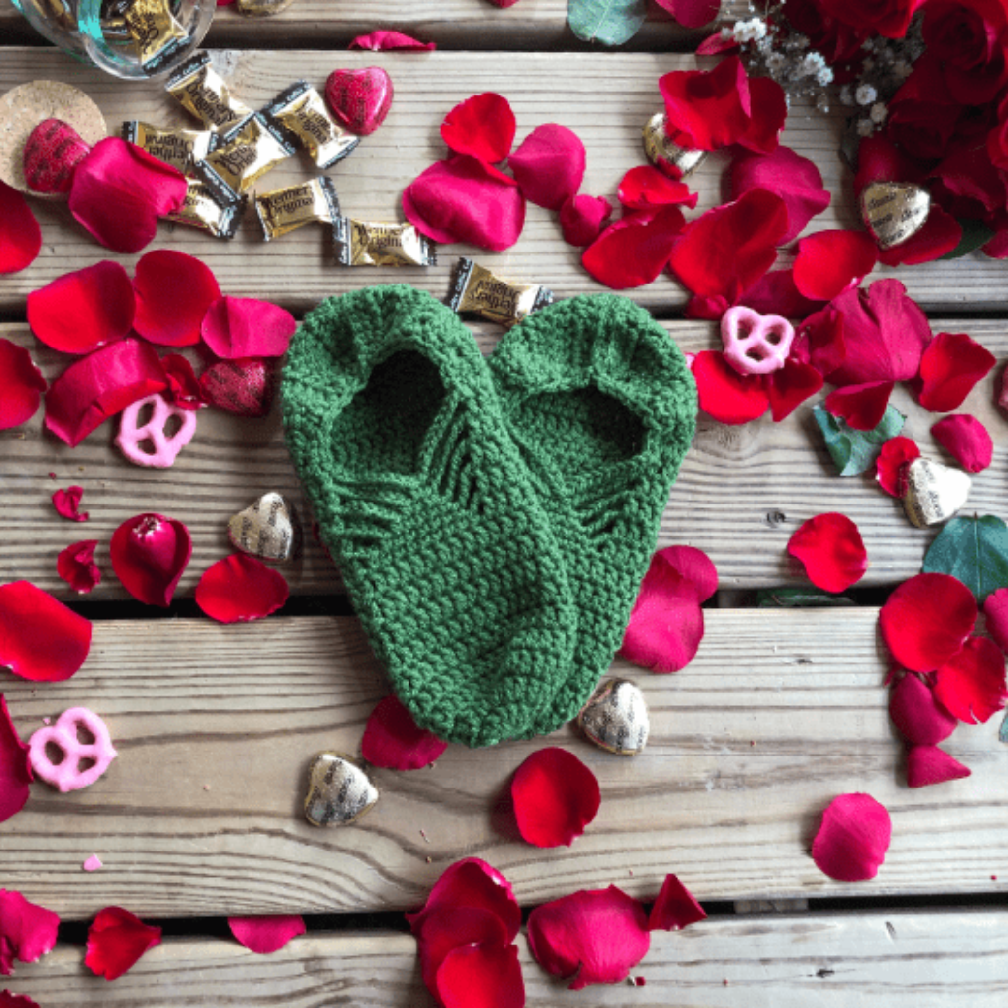 A pair of green slippers are laid out on a background of wooden slats, rose petals, chocolate hearts, and caramel candies.