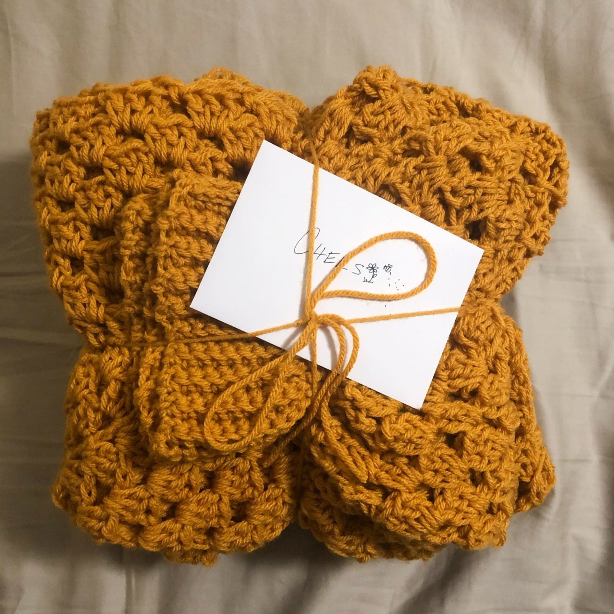 A Kelsie cardigan in the color Mustard is folded and packaged with a card that reads "Chels". The cardigan is laying on a neutral background. 