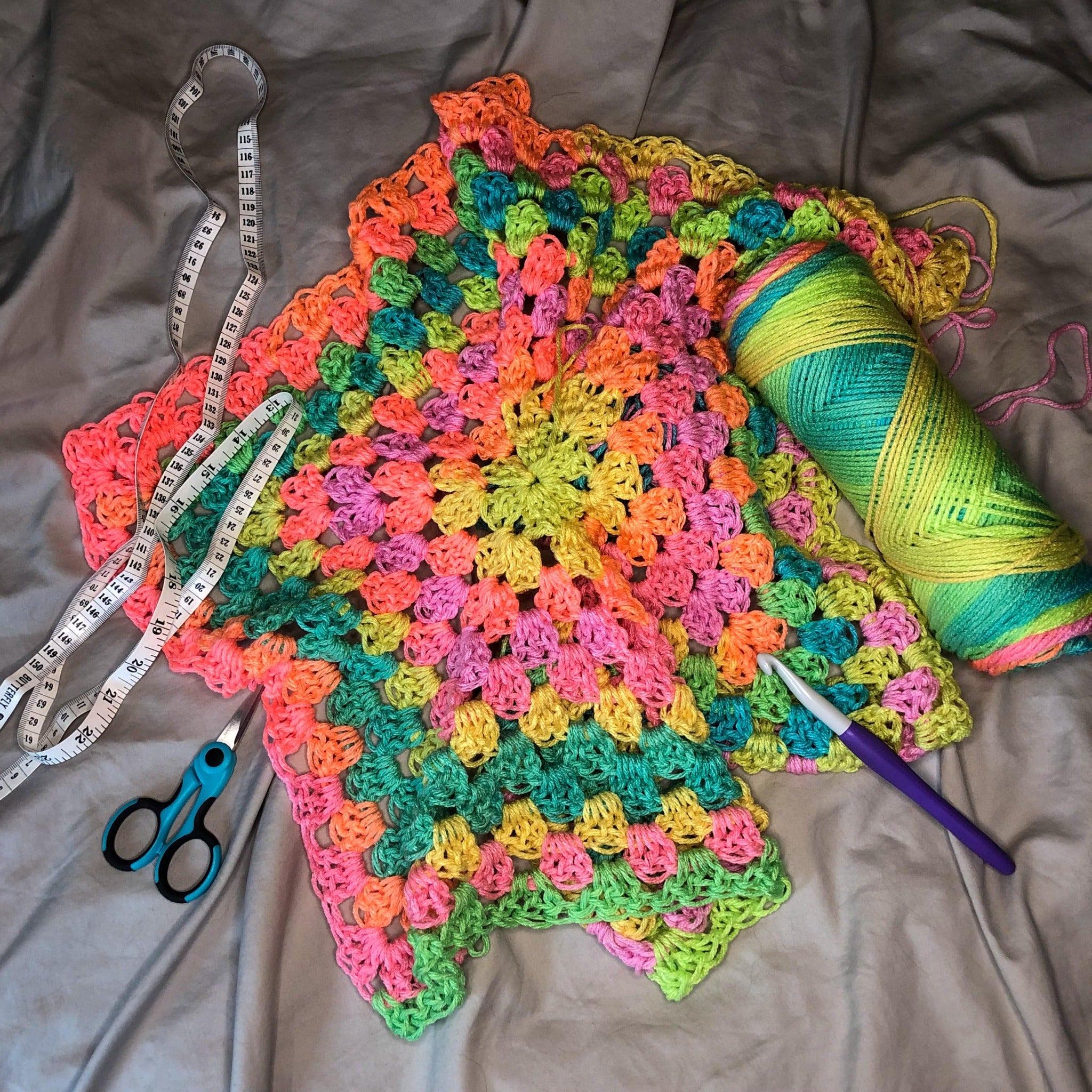 A crochet hexagon is laid out next to a skein of yarn in the colorway Retro Stripe with a crochet hook, blue scissors, and a measuring tape on a neutral background.