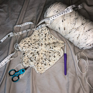 A crochet hexagon is laid out next to a skein of yarn in the colorway Pearl Fleck with a crochet hook, blue scissors, and a measuring tape on a neutral background.