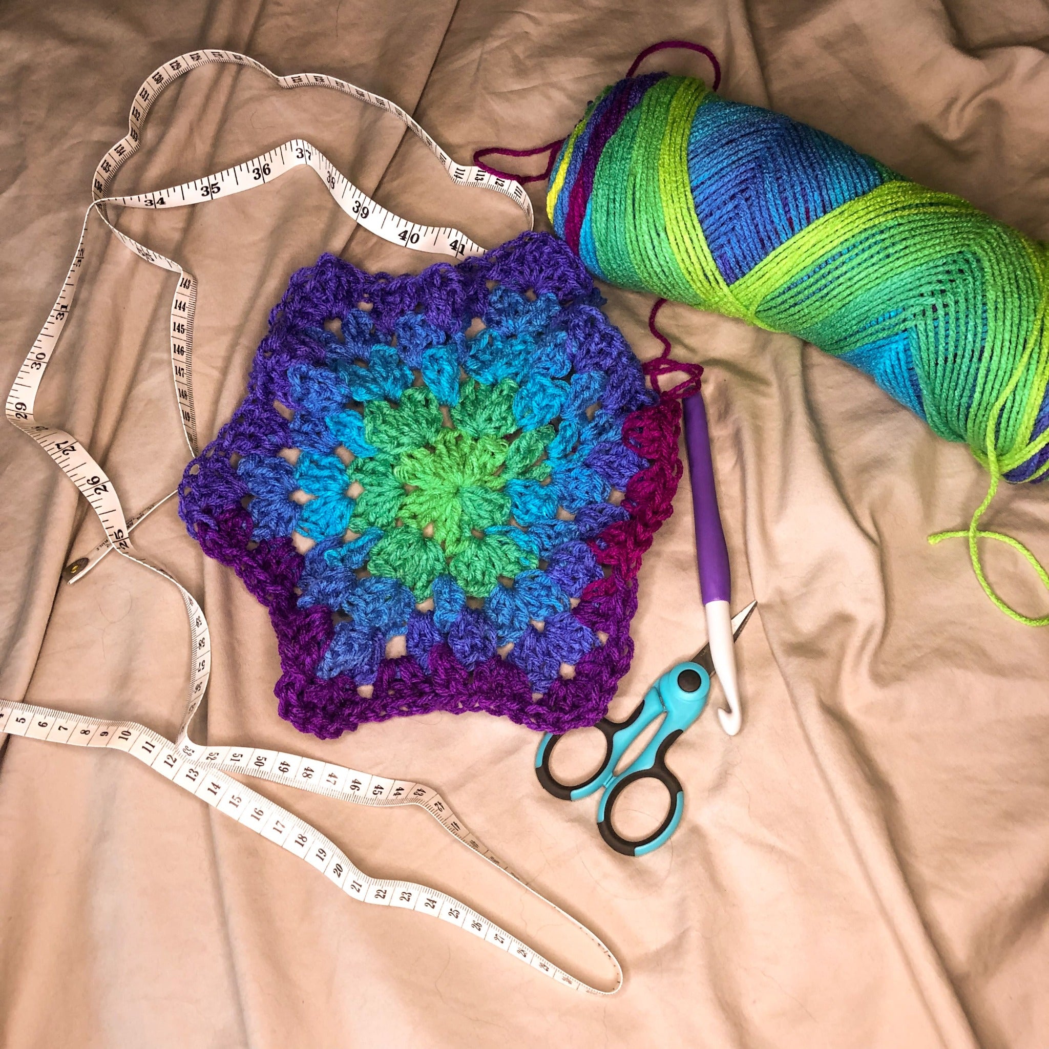 A crochet hexagon is laid out next to a skein of yarn in the colorway Parrot Stripe with a crochet hook, blue scissors, and a measuring tape on a neutral background.