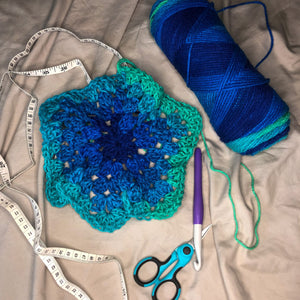 A crochet hexagon is laid out next to a skein of yarn in the colorway Cool Stripe with a crochet hook, blue scissors, and a measuring tape on a neutral background. 