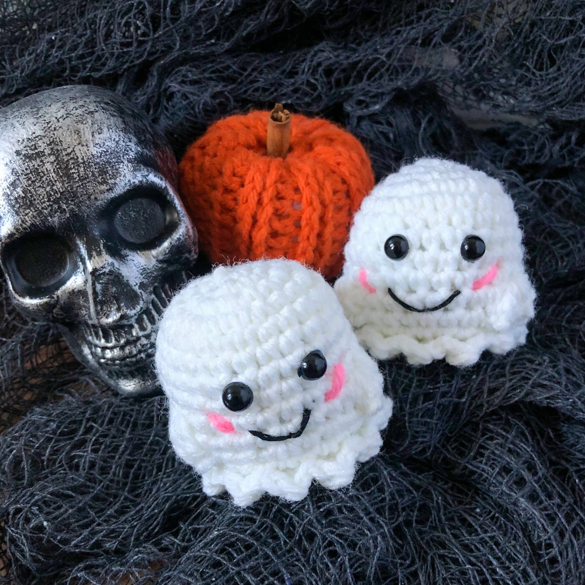 Two ghost plushies face the camera sitting with a mini pumpkin and a plastic, distressed skull Halloween decoration. The background is textured black "creepy fabric".