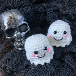 Two ghost plushies face the camera sitting with a plastic, distressed skull Halloween decoration. The background is textured black "creepy fabric".