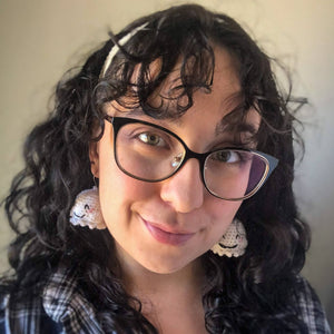 A woman with brown curly hair and glasses looks into the camera wearing ghost earrings and a black and white plaid flannel. 