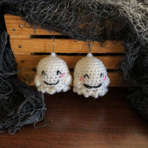 A pair of crochet ghost earrings face the camera with smiles hanging from a mini wooden crate with some creepy fabric around them. 