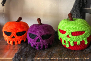 Three poison apples sit together on a shelf with creepy fabric behind and partially under two of the poison apples. The apples are crocheted with different colors and have poison dripping off them in the shape of skulls. 