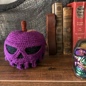 A crocheted poison apple with a black base and purple poison dripping down is on a bookshelf with some creepy fabric in the background and old books. The poison forms a scull as it is dripping.