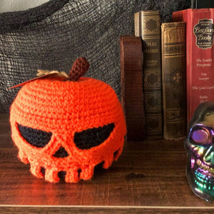 A crocheted poison apple with a black base and orange poison dripping down is on a bookshelf with some creepy fabric in the background and old books. The poison forms a scull as it is dripping.