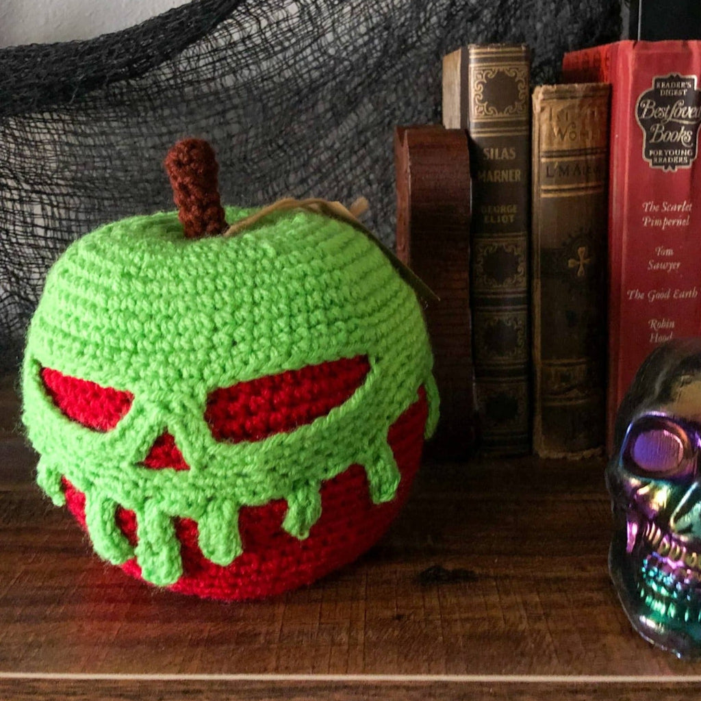 A crocheted poison apple with a red base and green poison dripping down is on a bookshelf with some creepy fabric in the background and old books . The poison forms a scull as it is dripping.