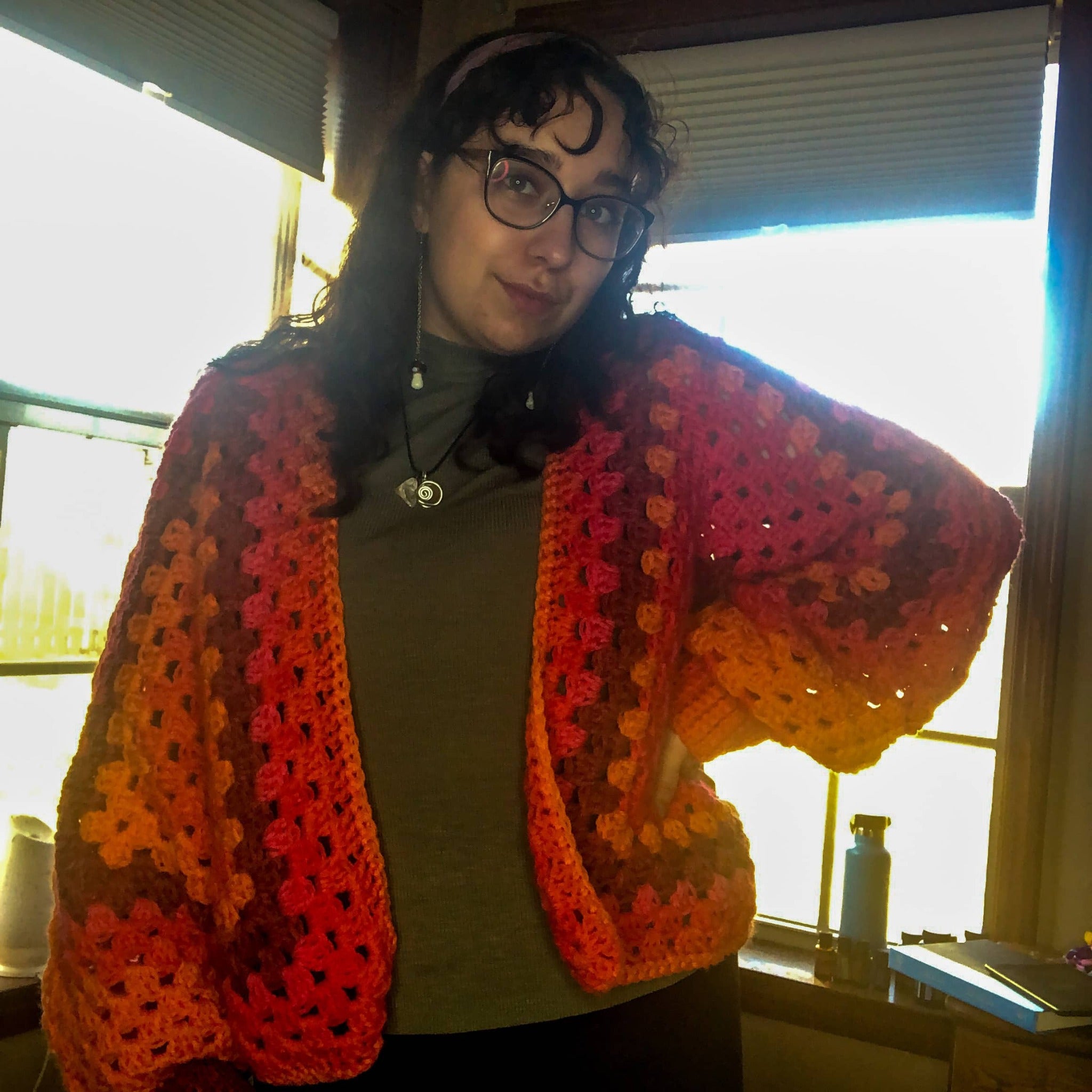 A woman with brown curly hair is wearing a cropped Kelsie cardigan in the colorway Scarlet Sizzle (a blend of burgundy, pink, and orange), facing the camera. She is also wearing a headband, glasses, drop mushroom earrings, and a green turtleneck. 