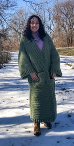 A woman with brown curly hair stands facing the camera and smiling with her hands in her pockets. She is standing in the snow and wearing a giant, thick, sage green ankle duster cardigan.