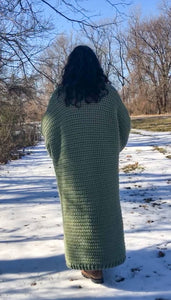 A woman with brown curly hair stands with her back to the camera and her arms in front of her. She is standing in the snow and wearing a giant, thick, sage green ankle duster cardigan.