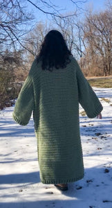 A woman with brown curly hair stands with her back to the camera and arms by her sides. She is standing in the snow and wearing a giant, thick, sage green ankle duster cardigan.