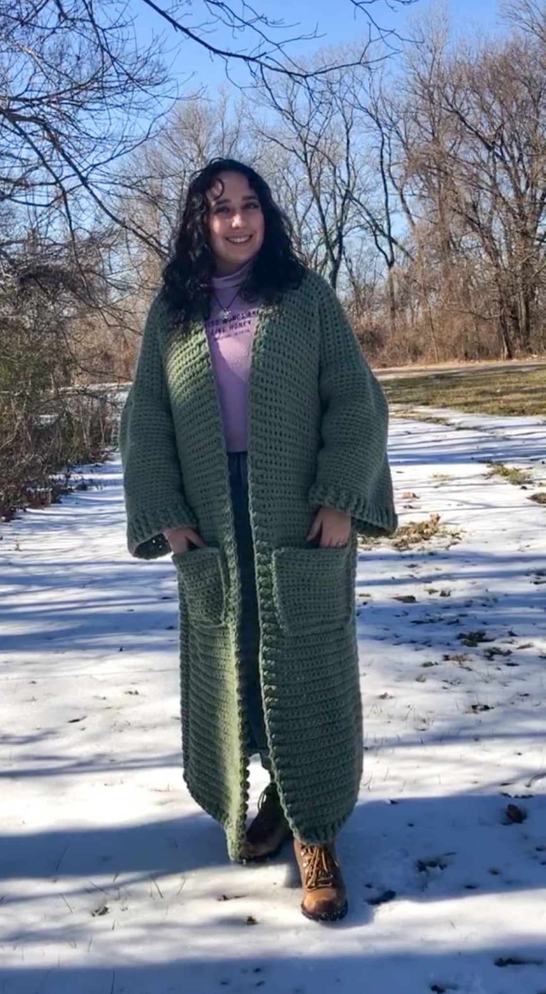 A woman with brown curly hair stands facing the camera and smiling with her hands in her pockets. She is standing in the snow and wearing a giant, thick, sage green ankle duster cardigan.