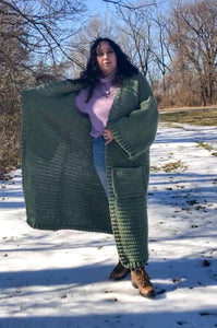 A woman with brown curly hair stands facing the camera wearing a purple turtleneck, jeans, and a giant sage green ankle duster cardigan. She is standing with one foot forward and one arm outstretched to show the inside lining of the ankle duster. 