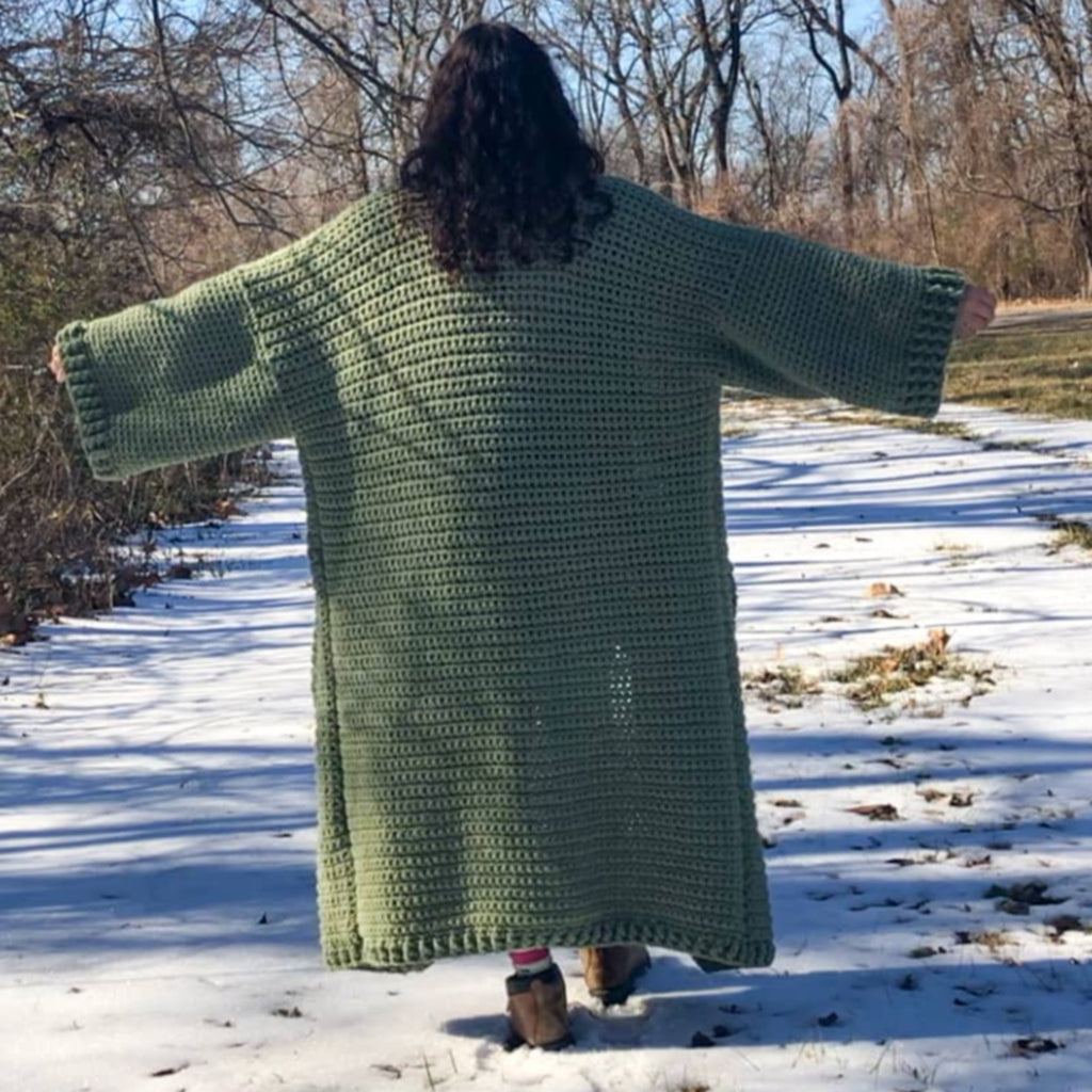 A woman with brown curly hair stands with her back to the camera and arms stretched out. She is standing in the snow and wearing a giant, thick, sage green ankle duster cardigan.