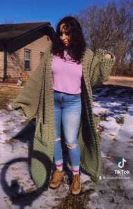 A woman with brown curly hair dances with one arm up, the other pointing down, and feet slightly apart. She is wearing a purple turtleneck, jeans, boots, and a sage green ankle duster cardigan. 