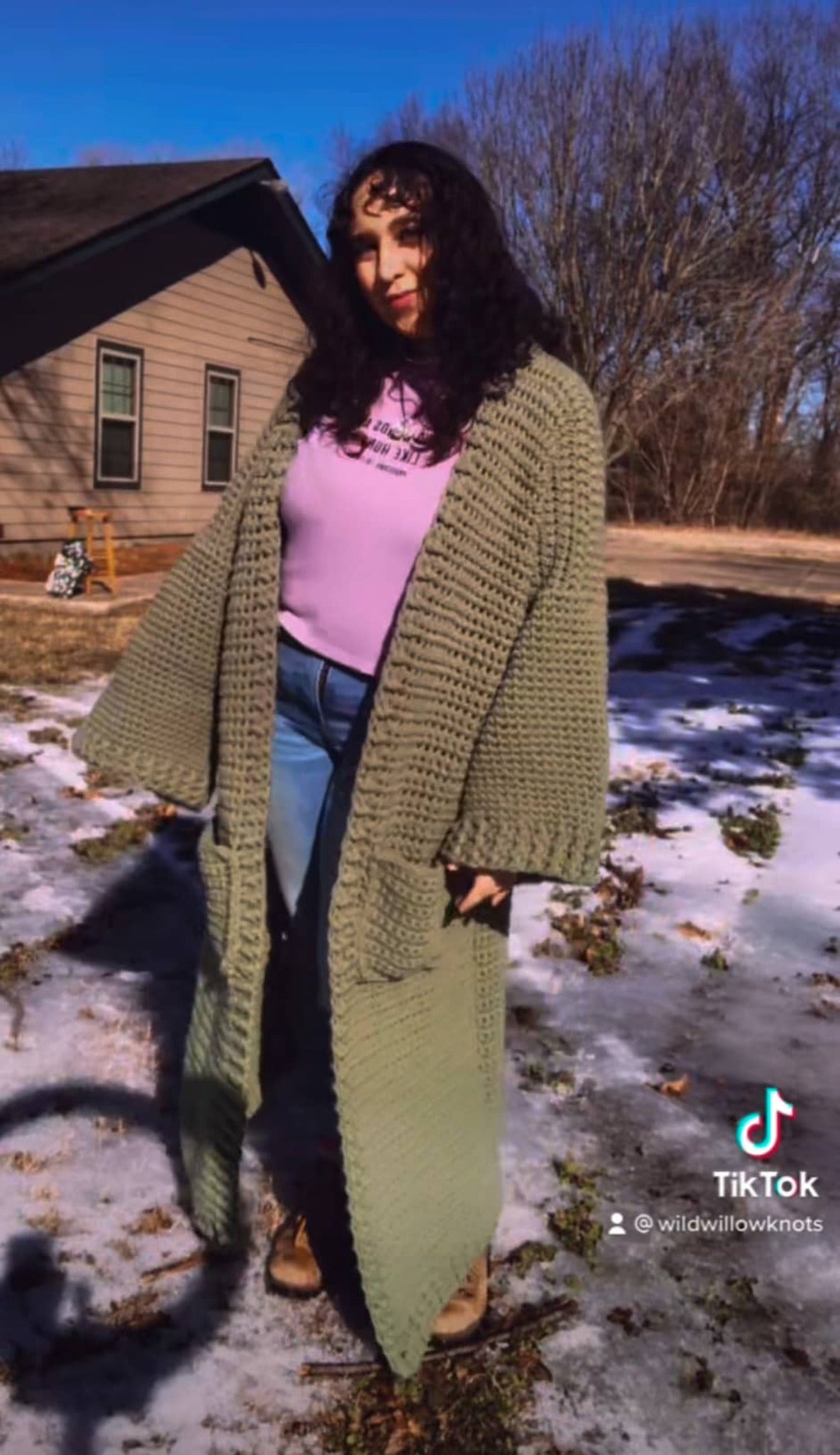 A woman with curly brown hair is standing facing the camera with her body tilted to the side as if she is swaying. She is wearing a purple turtleneck, jeans, boots, and a sage green ankle duster cardigan. 