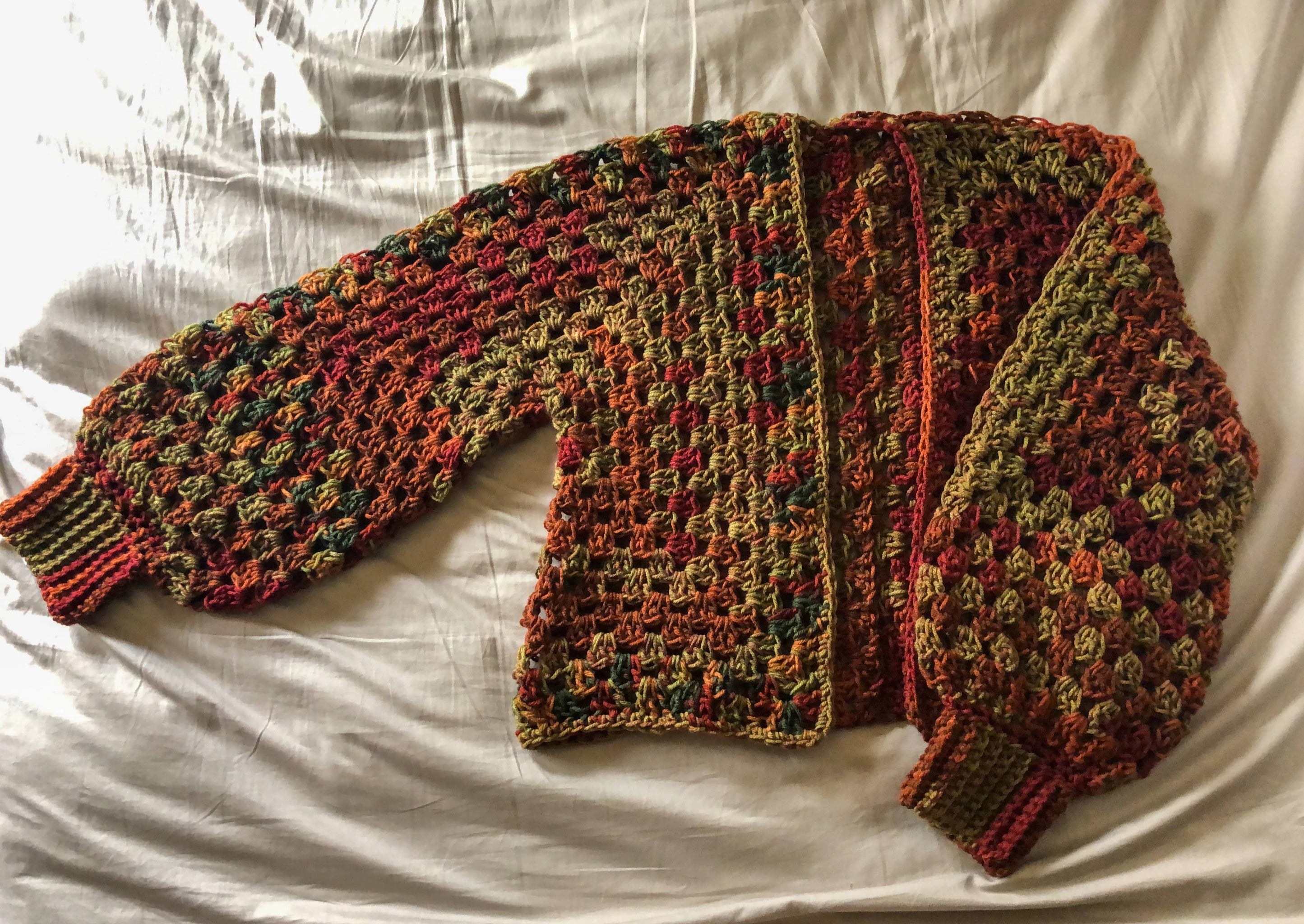 A Kelsie cardigan in the colorway Autumn Stripe (a blend of reds, greens, oranges, and browns) is laid out on a neutral background with one sleeve outstretched.