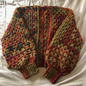 A cropped Kelsie cardigan in a the colorway of autumn stripe (a blend of reds, oranges, greens, and browns) is laid out on a neutral background.