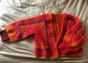 A Kelsie cardigan in the colorway Scarlet Sizzle (a blend of burgundy, pink, and orange) is laid out on a neutral background with one sleeve outstretched. 