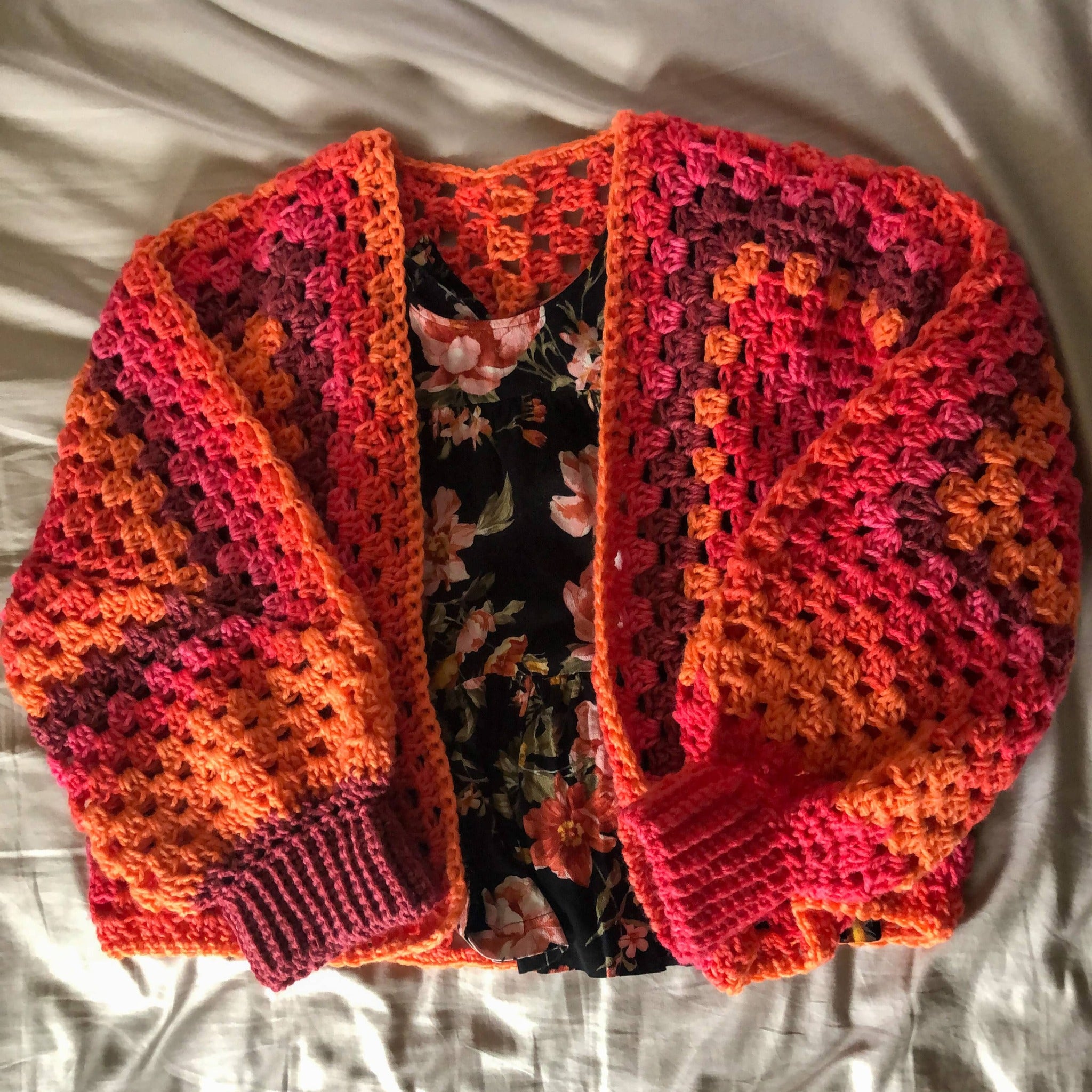 A cardigan in the colorway Scarlet Sizzle (a blend of burgundy, pink, and orange) is laying on a neutral background with a black shirt inside with a flower design. 