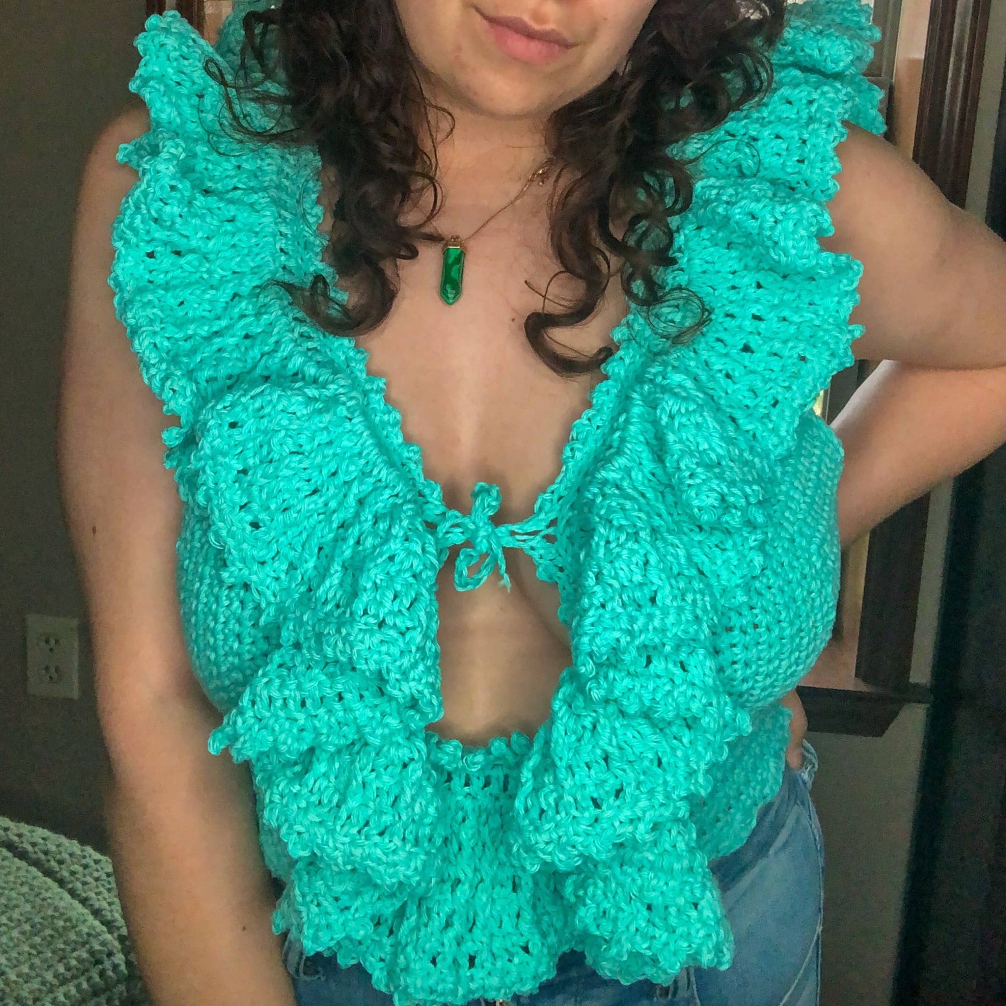 A woman with brown curly hair is wearing a teal, frilly crochet top. She has one hand on her hip and is facing the camera. 