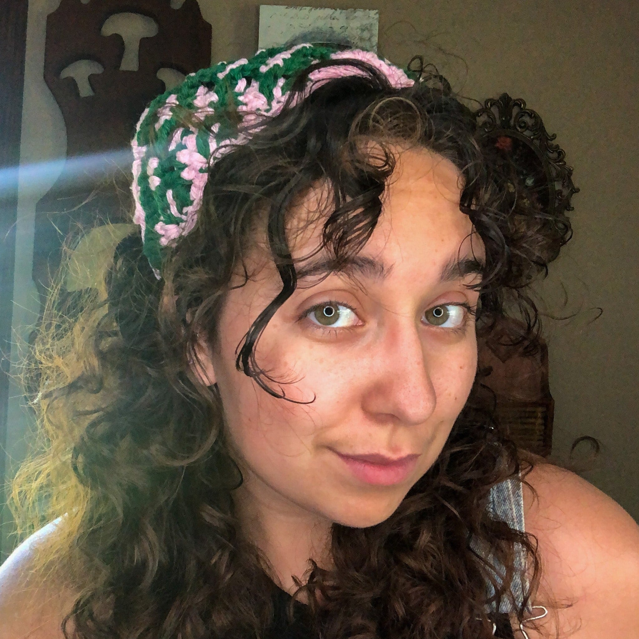 A woman with brown curly hair looks into the camera wearing a green and pink head scarf bandana.