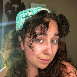 A woman with brown curly hair looks into the camera wearing a teal and pink head scarf bandana.