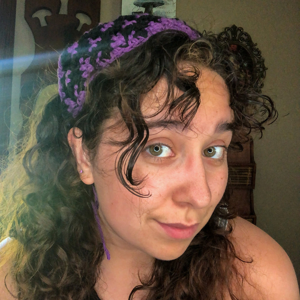 A woman with brown curly hair looks into the camera wearing a purple and black head scarf bandana.