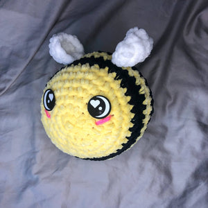 A giant plushie bee made of chenille yarn and with felt eyes faces to the left of the camera on a neutral background. 