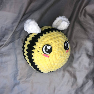 A giant plushie bee made of chenille yarn with big felt eyes faces to the right of the camera on a neutral background. 