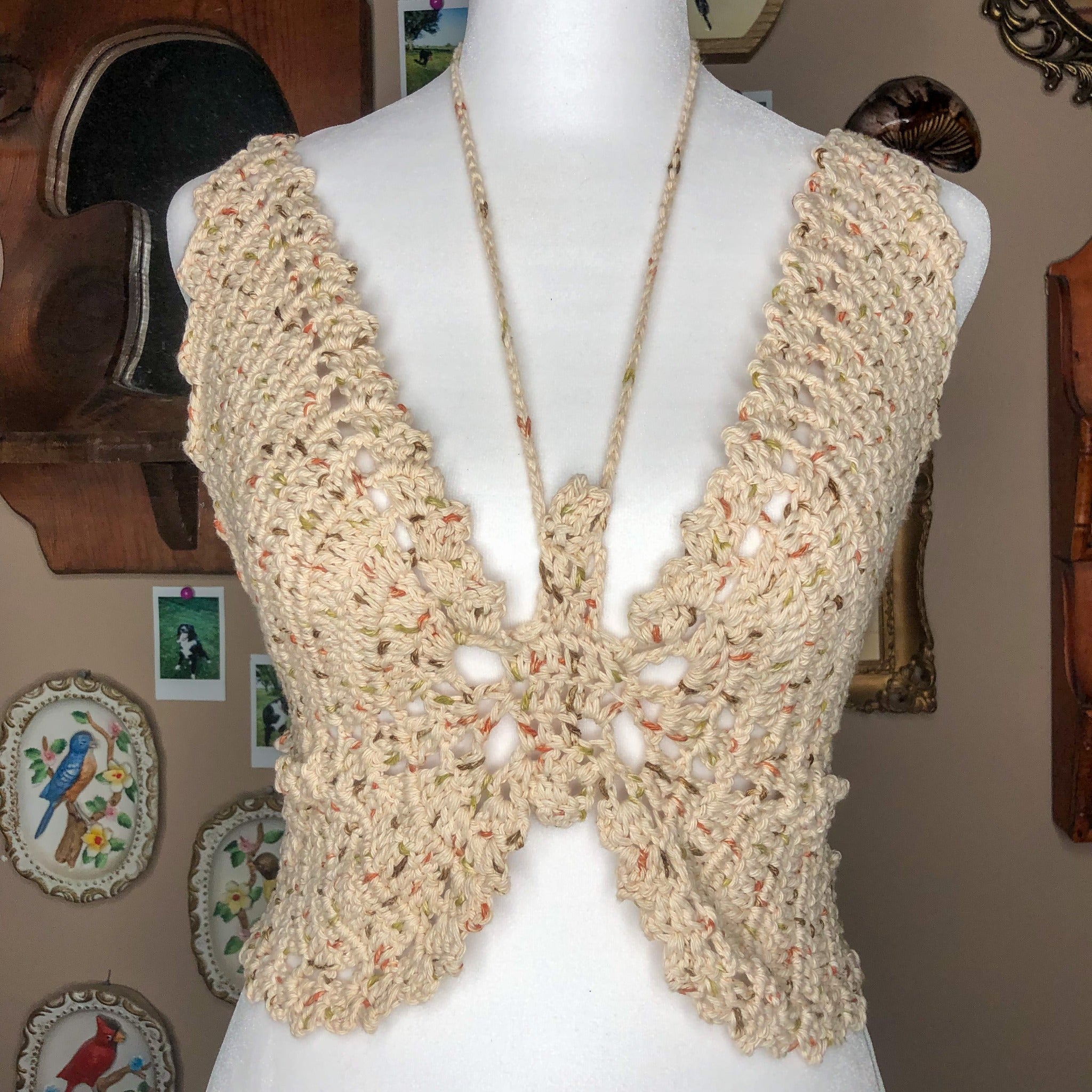 A crochet butterfly-shaped top is on a mannequin, facing the camera straight on. There are a few trinkets in the background such as a mushroom mirror, a few framed porcelain birds, and some frames.