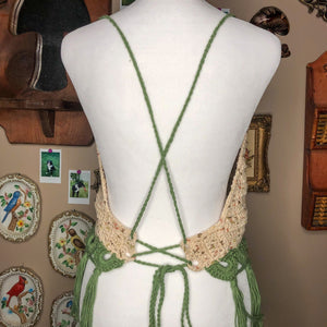 A sage top is on a mannequin, facing away from the camera to show off the crisscross straps. In the background are various wall hanging decorations including a mushroom mirror and porcelain birds. 