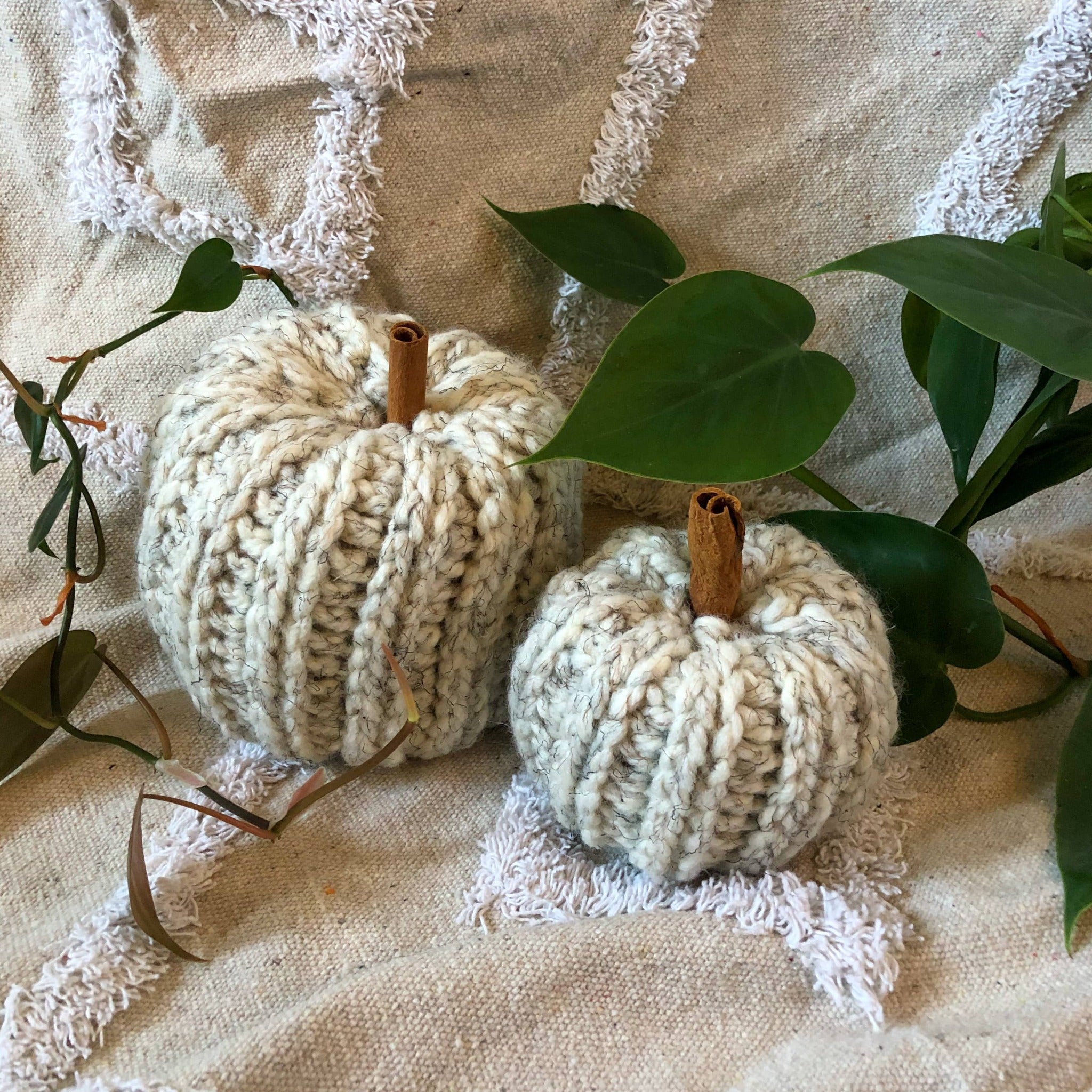 Two marbled white knit pumpkins with cinnamon sticks for stems sit on a rug with plant vines around them. 