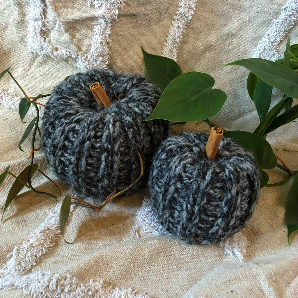 Two knit pumpkins made with charcoal gray marbled yarn and cinnamon sticks for the stem are placed on a rug with a plant vine around them.