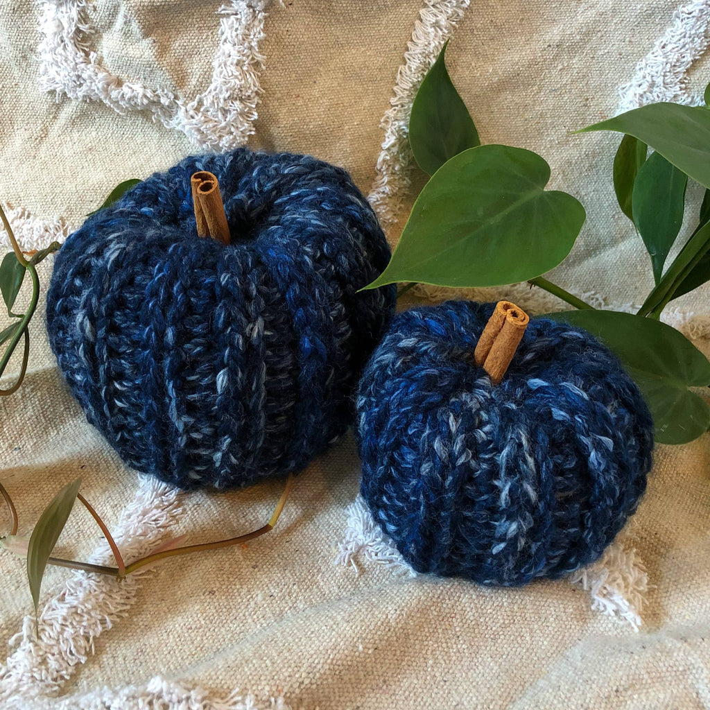 Two marbled blue knit pumpkins with cinnamon sticks for stems sit on a rug with plant vines around them. 