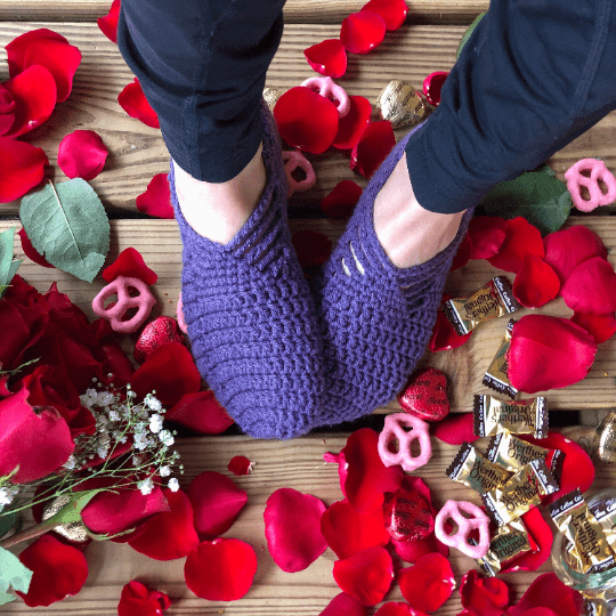 A woman wearing black leggings is wearing a pair of purple house slippers on a background of rose petals, pink yogurt covered pretzels, and caramel candies. 