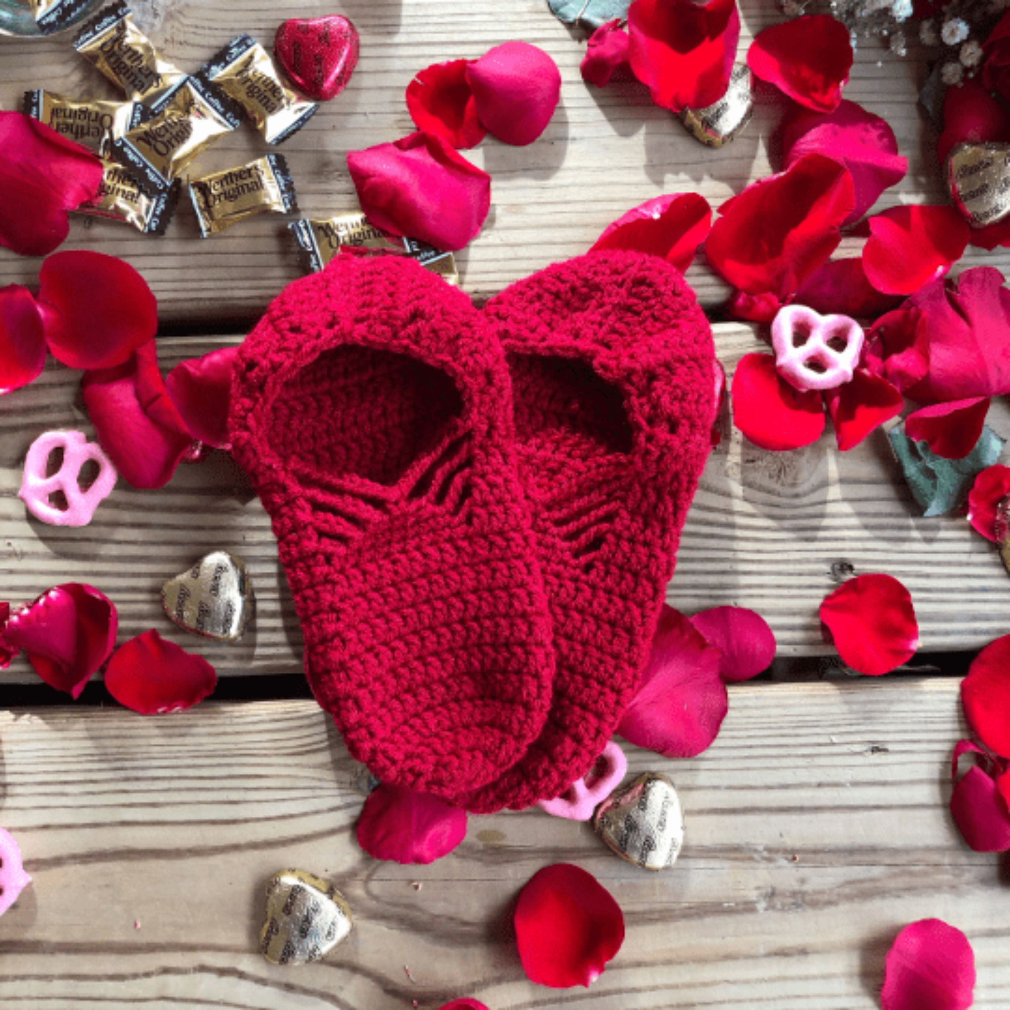 A pair of red slippers are laid out on a background of wooden slats, rose petals, chocolate hearts, and caramel candies.