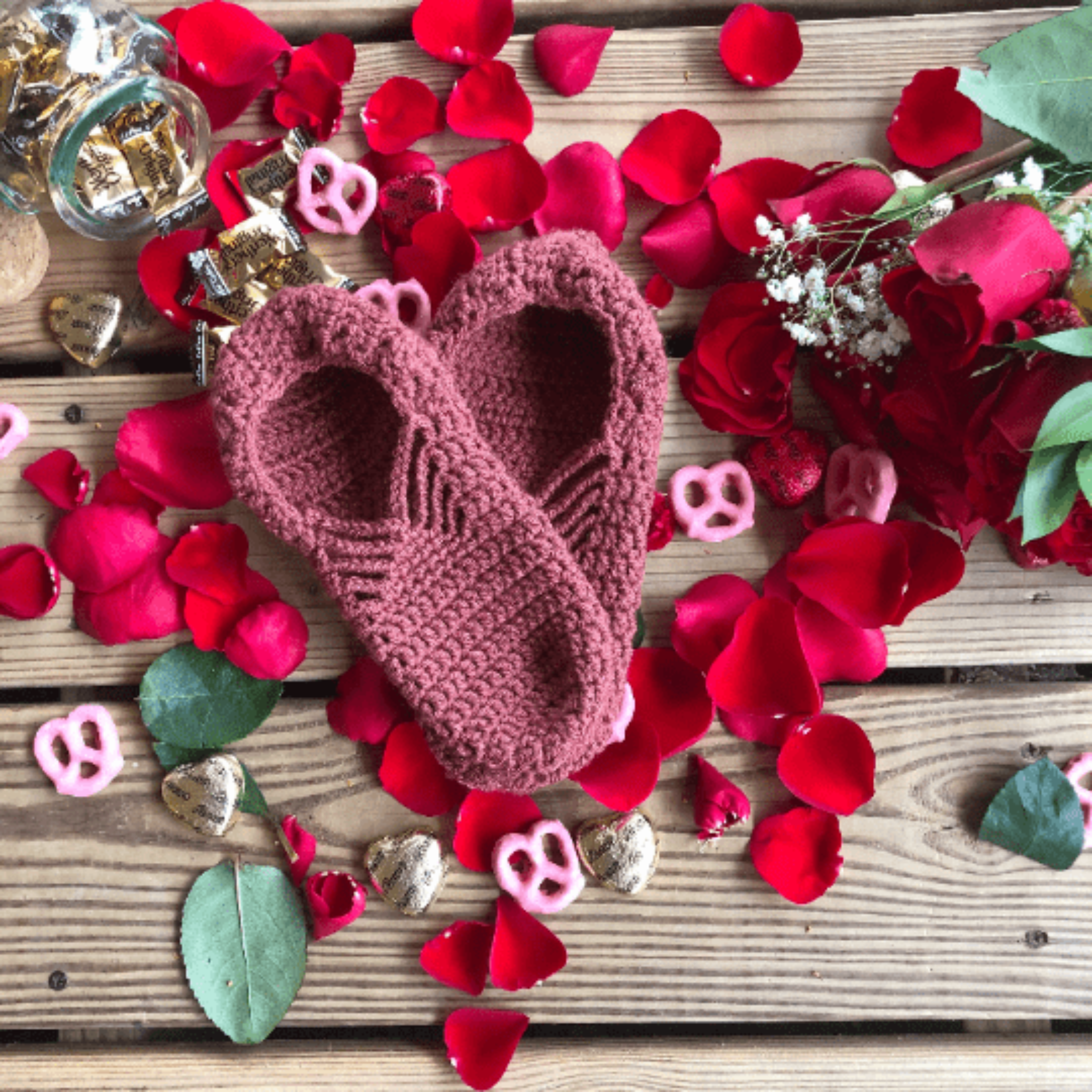 A pair of pink spice slippers are laid out on a background of wooden slats, rose petals, chocolate hearts, and caramel candies.